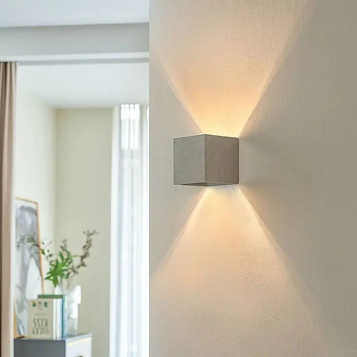 Lindby Wandleuchte Jayden - L x B x H: 11,5 x 11,5 x 11,5 cm, alu / grau / zink, G9 - Eine graue, würfelförmige Wandlampe ist an einer hellen Wand montiert und wirft warmes Licht nach oben und unten in einem modernen Raum.