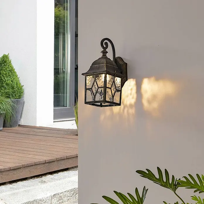 Lindby Wandleuchte Purnima - L x B x H: 17,8 x 15,1 x 31,8 cm, bronze / altmessing, bronze / altmessing, E27 - Antike Messing-Außenwandlaterne mit gemustertem Glas, beleuchtet eine Terrassenwand.