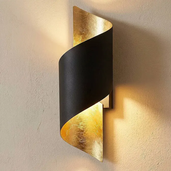 Lindby Wandleuchte Desirio - schwarz, schwarz, E14 - Schwarze und Blattgold-Spirale-Wandleuchte wirft warmes Licht nach oben und unten an eine strukturierte Wand.