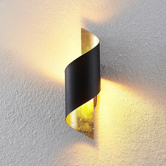 Lindby Wandleuchte Desirio - L x B x H: 13 x 10,5 x 35 cm, schwarz, schwarz, E14 - Eine schwarz-goldene spiralförmige Wandleuchte ist an einer strukturierten Wand montiert und wirft warmes Licht nach oben und unten.