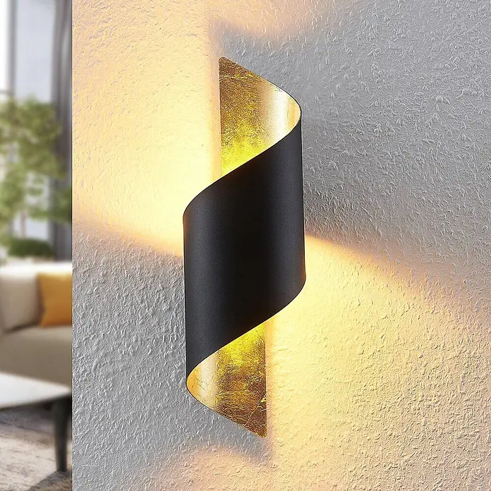 Lindby Wandleuchte Desirio - schwarz, schwarz, E14 - Moderne schwarz-goldene gedrehte Wandleuchte, die warmes Licht nach oben und unten auf eine strukturierte Wand in einem Wohnzimmer wirft.