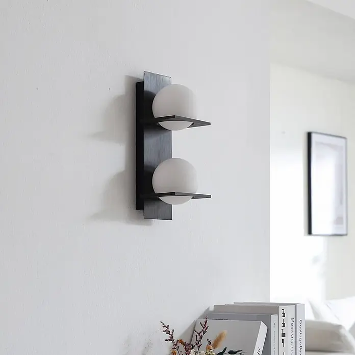 Lucande Wandleuchte Kulka - L x B x H: 13 x 13 x 32 cm, schwarz, G9 - Moderne Wandleuchte mit dunklem, rechteckigem Sockel und zwei weißen Kugeldiffusoren aus Glas, an einer Wand.