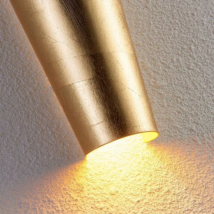 Lindby Wandleuchte Conan - messing / gold, messing / gold, G9 - Goldene Wandlampe wirft warmes Licht auf eine strukturierte Wand.