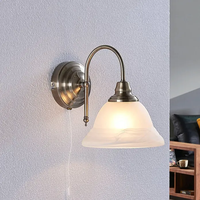 Lindby Wandleuchte Hanna - bronze / altmessing, weiß / opal, E14 - Wandleuchte aus Altmessing mit mattem Glasschirm und Zugschalter an einer strukturierten Wand.