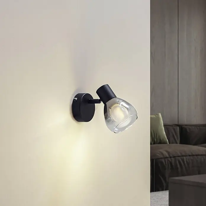 Lindby Wandleuchte Pendura - L x B x H: 9 x 9 x 12,5 cm, schwarz, alu / grau / zink, E14 - Eine schwarze Wandleuchte mit klarem Glasschirm spendet warmes Licht an einer hellen Wand, neben einem Sofa.