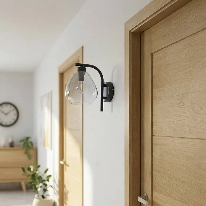 Lindby Wandleuchte Pendura - schwarz, alu / grau / zink, E14 - Schwarze Metall-Wandleuchte mit klarem Glasschirm, an einer weißen Wand zwischen zwei hellen Holztüren montiert.