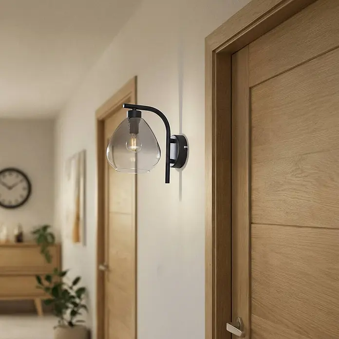 Lindby Wandleuchte Pendura - schwarz, alu / grau / zink, E14 - Moderne schwarze Wandleuchte mit rauchigem Glasschirm beleuchtet einen Flur mit Holztüren. Die freiliegende Glühbirne wirkt vintage.