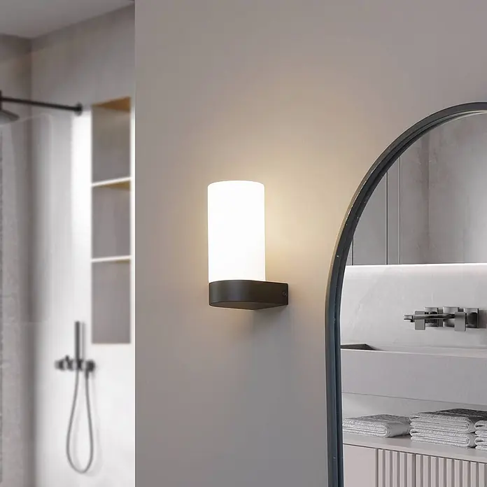 Lucande Wandleuchte Basolo - weiß / opal, E14 - Wandmontierte zylindrische Leuchte mit dunklem Sockel und weißem Diffusor, die ein modernes Badezimmer beleuchtet.