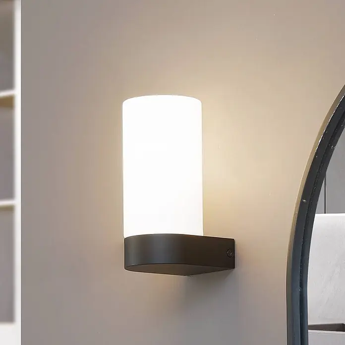 Lucande Wandleuchte Basolo - weiß / opal, E14 - Eine moderne Wandleuchte mit dunklem Sockel und weißem Zylinderdiffusor beleuchtet eine Wand neben einem Spiegel.