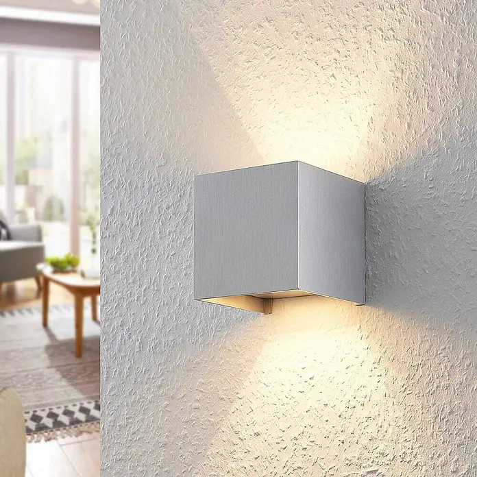 Arcchio Wandleuchte Zuzana - alu / grau / zink, G9 - Moderne silbergraue würfelförmige Wandleuchte, die eine strukturierte weiße Wand mit Ober- und Unterlicht in einem Wohnzimmer beleuchtet.