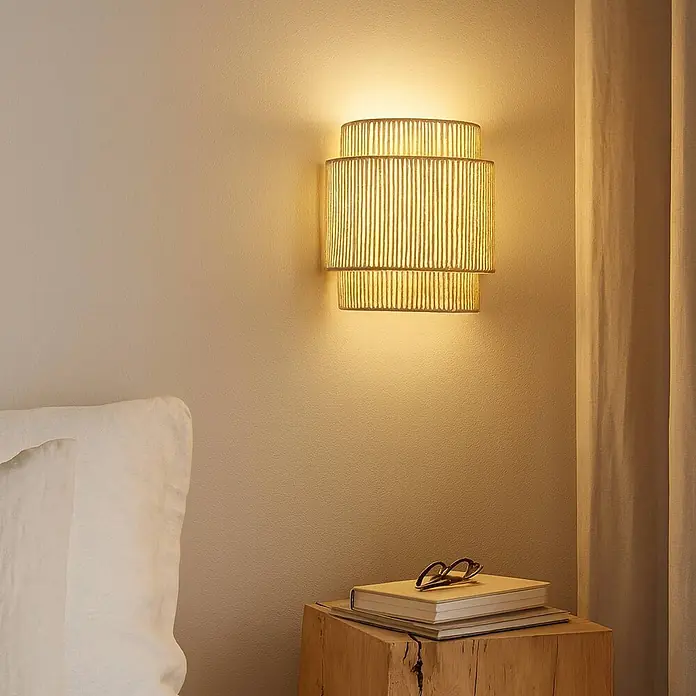Lindby Wandleuchte Anuva - L x B x H: 13 x 22 x 25 cm, creme / amber, E27 - Wandleuchte aus Naturfaser mit vertikaler Textur, die ein warmes Licht in einem Schlafzimmer wirft.