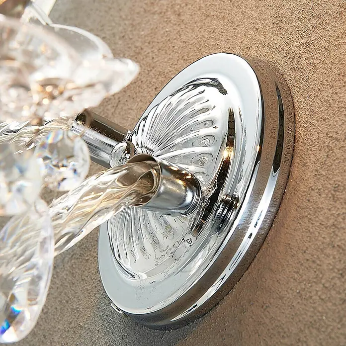 Lindby Wandleuchte Merida - L x B x H: 18,5 x 33 x 35 cm, chrom / silber, klar / transparent, E14 - Verchromte Wandleuchtenbasis mit gedrehten klaren Kristallakzenten, an einer strukturierten beigen Wand montiert.