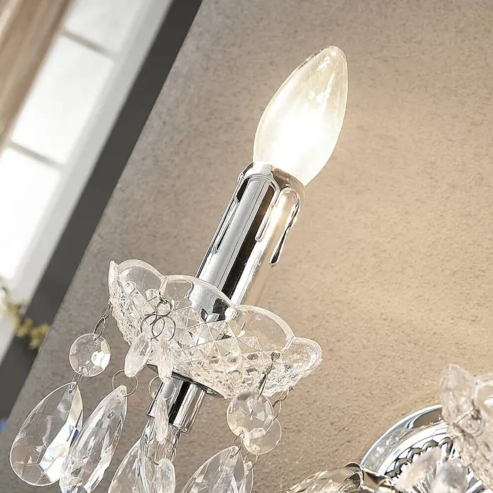 Lindby Wandleuchte Merida - L x B x H: 18,5 x 33 x 35 cm, chrom / silber, klar / transparent, E14 - Verchromte Wandleuchte mit beleuchteter Kerzenlampe und funkelnden klaren Kristallanhängern, an einer strukturierten Wand montiert.