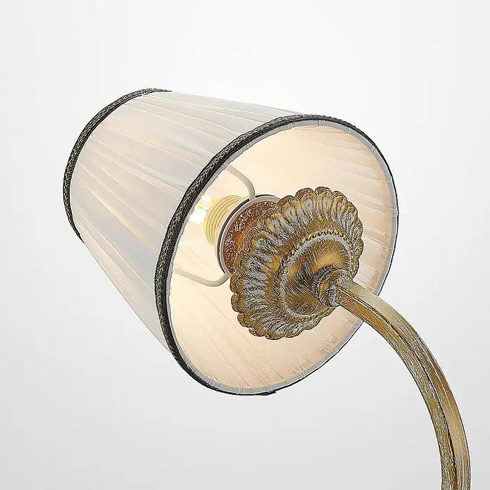 Lindby Wandleuchte Lumiel - bronze / altmessing, creme / amber, E14 - Nahaufnahme eines weißen plissierten Lampenschirms mit dunkler Borte, der eine antikgoldene Lampenfassung und Glühbirne zeigt.