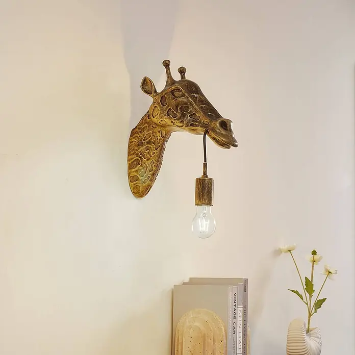 Lindby Wandleuchte Nirvathia - bronze / altmessing, E27 - Wandleuchte in Form eines goldfarbenen Giraffenkopfes mit sichtbarer Glühbirne, an einer hellen Wand über Büchern.