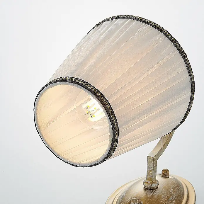Lindby Wandleuchte Lumiel - bronze / altmessing, creme / amber, E14 - Eine Lampe mit plissiertem, cremefarbenem Schirm, dunkler Flechtverzierung und sichtbarer LED-Birne auf einem goldfarbenen Sockel.