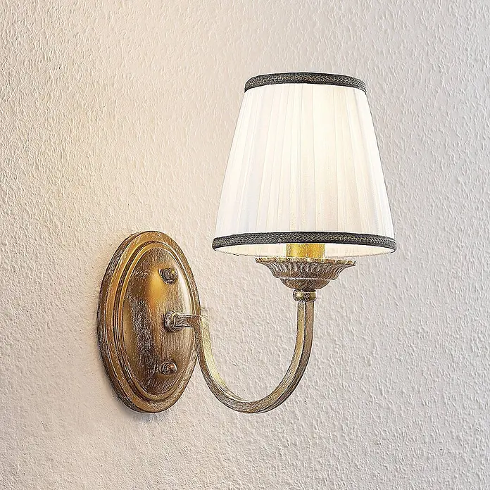 Lindby Wandleuchte Lumiel - bronze / altmessing, creme / amber, E14 - Wandleuchte aus antikem Messing mit plissiertem weißem Stoffschirm und dunklem Rand, beleuchtet und an einer strukturierten Wand montiert.