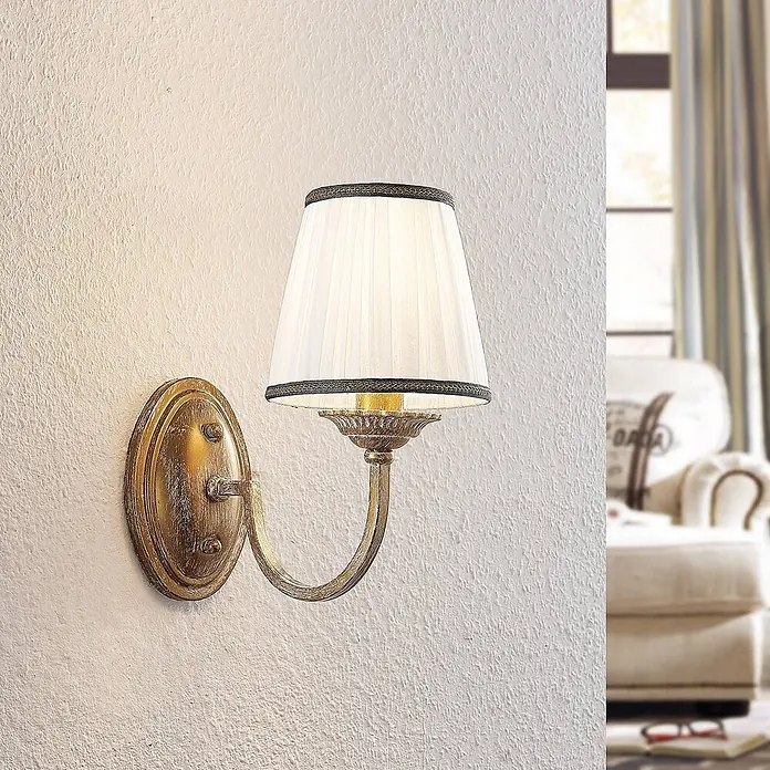 Lindby Wandleuchte Lumiel - bronze / altmessing, creme / amber, E14 - Elegante goldfarbene Wandleuchte mit plissiertem weißen Stoffschirm an einer strukturierten Wand.