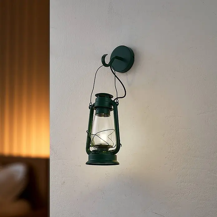 Lindby Wandleuchte Raisa - grün, E14 - Eine dunkelgrüne, an der Wand montierte elektrische Laterne mit einem hell leuchtenden Klarglasglobus, der eine strukturierte Wand beleuchtet.