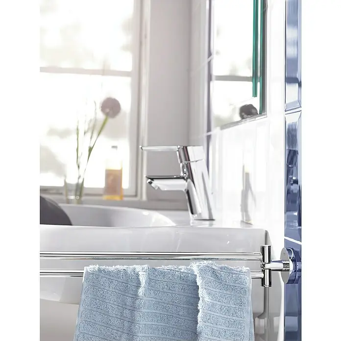 Grohe Essentials Handtuchhalter - 2-armig, Chrom, Glänzend - Ein hellblaues Handtuch hängt an einem Handtuchhalter vor einem Waschbecken und einem Chromwasserhahn in einem hellen Badezimmer.