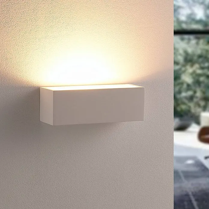 Lindby Wandleuchte Santino - L x B x H: 7,5 x 21,8 x 7,5 cm, weiß / opal, G9 - Weiße rechteckige Wandleuchte, die warmes Licht nach oben und unten auf eine strukturierte Wand strahlt, mit unscharfem Außenhintergrund.