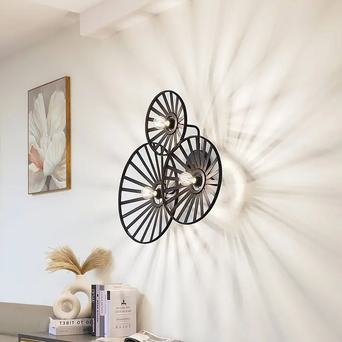 Lindby Wandleuchte Evandor - schwarz, E27 - Schwarze Wandleuchte aus Metall mit drei radförmigen Designs an einer Wand, die dekorative Schatten wirft.