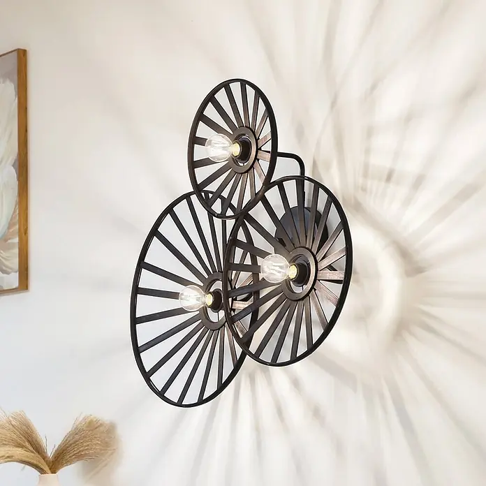 Lindby Wandleuchte Evandor - schwarz, E27 - Dunkle Metall-Wandleuchte mit drei radförmigen Rahmen und freiliegenden Glühbirnen, an einer Wand montiert und dekorative Schatten werfend.