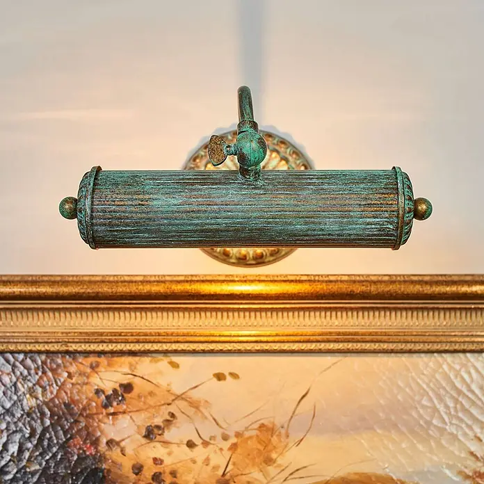 Lindby Wandleuchte Beno - bronze / altmessing, bronze / altmessing, E14 - Grünspan-Bilderleuchte beleuchtet ein goldgerahmtes Gemälde.