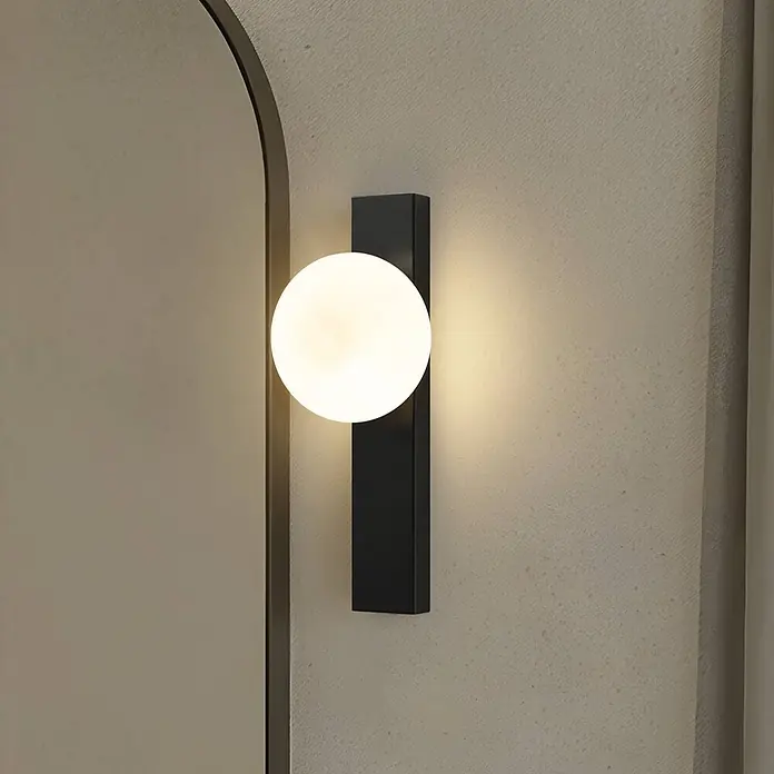 Lindby Wandleuchte Tavrik - schwarz, weiß / opal, G9 - Moderne schwarze, rechteckige Wandleuchte mit leuchtender Kugellampe, an einer strukturierten Wand neben einem gewölbten Rahmen montiert.