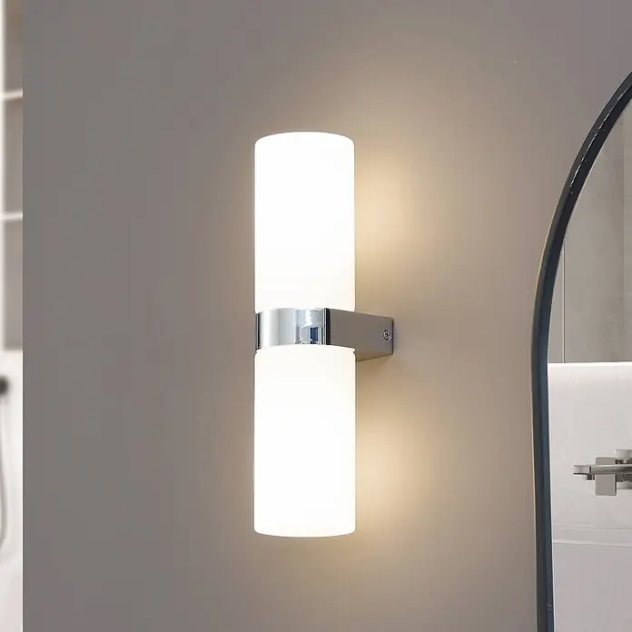Lucande Wandleuchte Basolo - L x B x H: 10 x 8 x 31,5 cm, chrom / silber, E14 - Wandleuchte mit zwei zylindrischen, mattierten Glasschirmen und Chromfuß, beleuchtet eine neutrale Wand neben einem Spiegel.