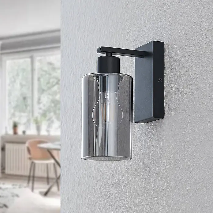 Lindby Wandleuchte Emavi - L x B x H: 15 x 10 x 23 cm, schwarz, schwarz, E27 - Schwarze Metall-Wandleuchte mit zylindrischem Rauchglas-Schirm und sichtbarer Glühbirne, an einer Wand montiert.