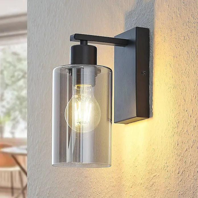 Lindby Wandleuchte Emavi - L x B x H: 15 x 10 x 23 cm, schwarz, schwarz, E27 - Moderne Wandleuchte aus schwarzem Metall und klarem Glaszylinder, an strukturierter Wand montiert und beleuchtet.
