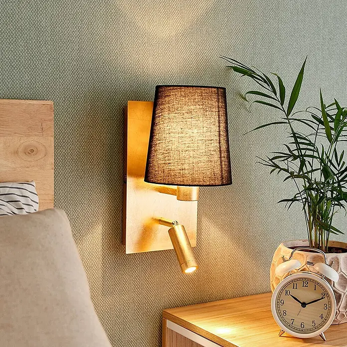 Lindby Wandleuchte Aiden - messing / gold, schwarz, E14 - Goldene Wandleuchte mit schwarzem Textilschirm und integriertem Lesespot, an einer strukturierten Wand neben einem Bett.
