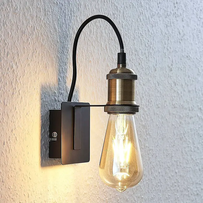 Lindby Wandleuchte Aurella - bronze / altmessing, E27 - Industrielle Wandleuchte mit dunklem Metallsockel, antikem Messing-Fassung und sichtbarer LED-Glühbirne an einer strukturierten Wand.