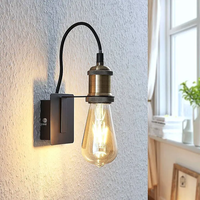 Lindby Wandleuchte Aurella - bronze / altmessing, E27 - Eine Wandleuchte im Industrial-Stil mit schwarzem Sockel, antikem Messingfassung und Vintage-LED-Birne, die eine strukturierte Wand beleuchtet.