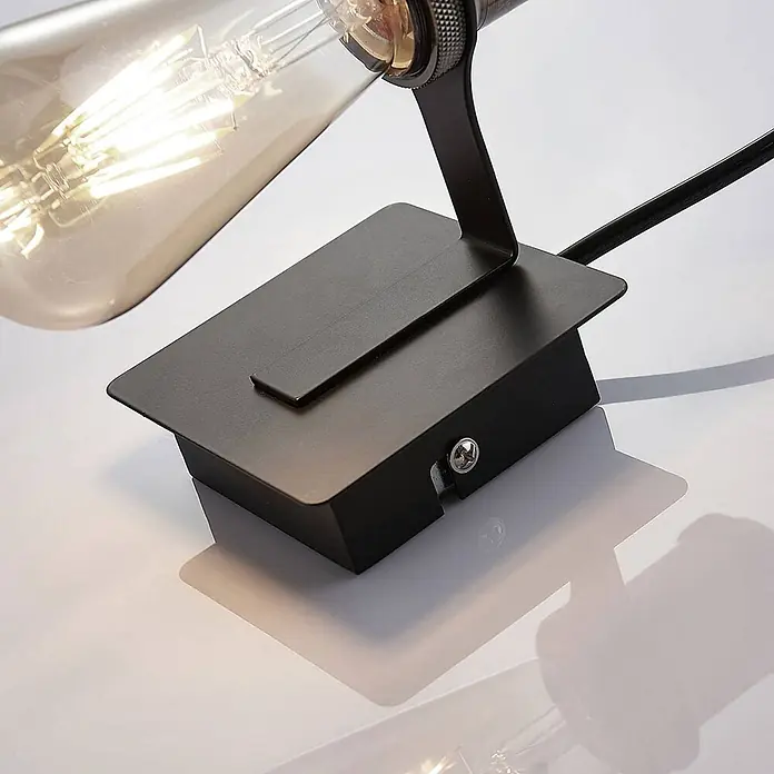 Lindby Wandleuchte Aurella - bronze / altmessing, E27 - Schwarzer Metallsockel einer Lampe mit sichtbarer Schraube, schwarzem Kabel und einem leuchtenden Edison-Glühbirnenteil.