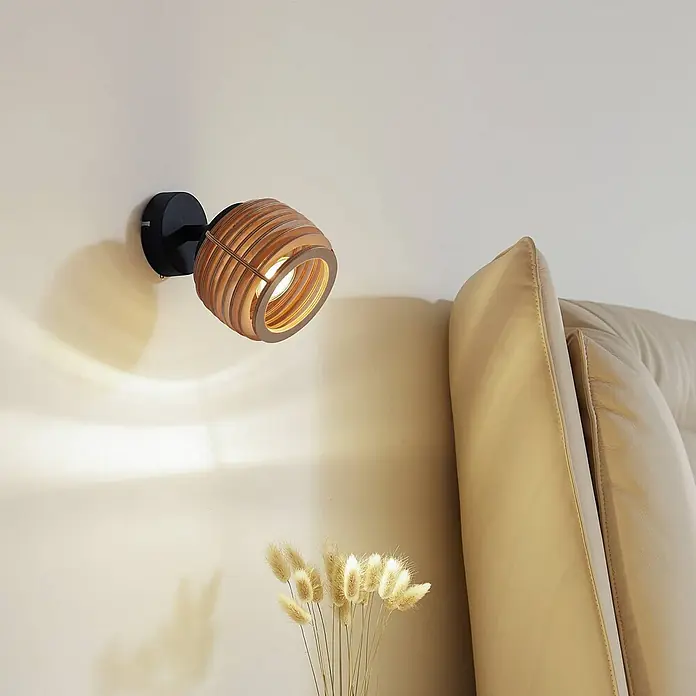 Lindby Wandleuchte Ediz - L x B x H: 17,3 x 12,7 x 12,7 cm, schwarz, Holz hell, GU10 - Wandstrahler mit Holzlamellenschirm, beleuchtet eine cremefarbene Wand neben einem beigen Sofa.