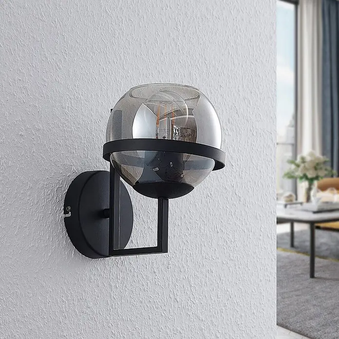 Lindby Wandleuchte Marinka - schwarz, klar / transparent, E27 - Moderne schwarze Wandleuchte mit Rauchglaskugel und sichtbarer Glühbirne, an einer strukturierten weißen Wand.