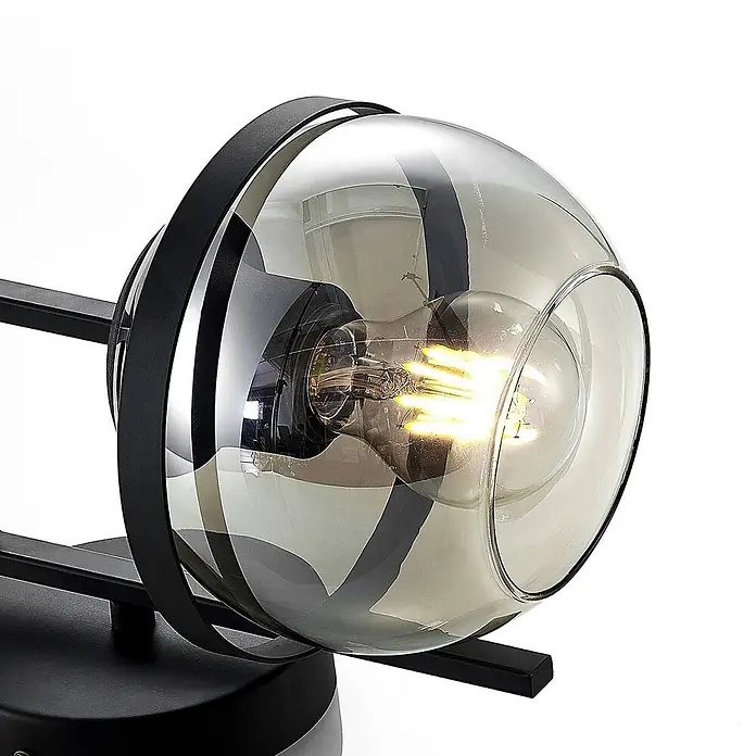 Lindby Wandleuchte Marinka - schwarz, klar / transparent, E27 - Moderne Lampe mit leuchtender Glühbirne in kugelförmigem Rauchglas-Schirm, gehalten von einem schwarzen Metallrahmen.
