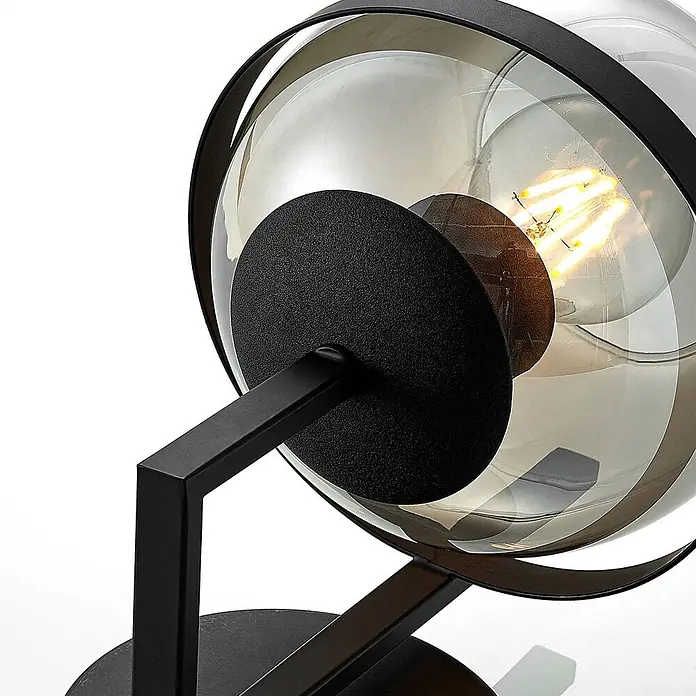 Lindby Wandleuchte Marinka - schwarz, klar / transparent, E27 - Moderne Lampe mit schwarzem Metallrahmen und rauchfarbenem Glaskugelschirm, der eine Edison-Glühbirne zeigt.