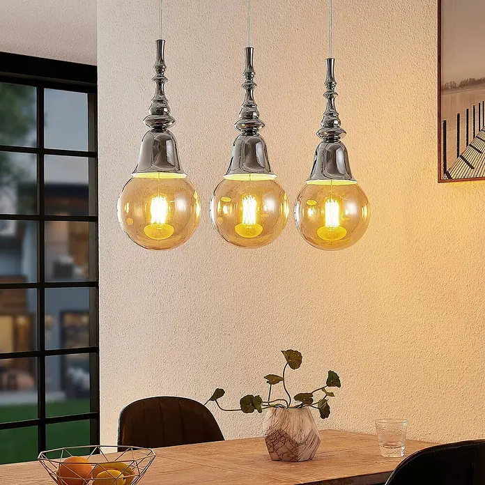 Lucande Pendelleuchte Gesja - L x B x H: 63,5 x 13,5 x 32,5 cm, chrom, E27 - Drei Pendelleuchten mit Chromakzenten und bernsteinfarbenen Glaskugeln beleuchten einen Esstisch aus Holz.