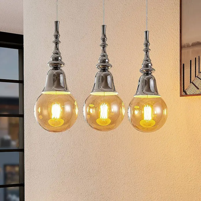 Lucande Pendelleuchte Gesja - L x B x H: 63,5 x 13,5 x 32,5 cm, chrom, E27 - Drei Pendelleuchten mit Chromfassungen und bernsteinfarbenen Glaskugeln beleuchten eine Wand.