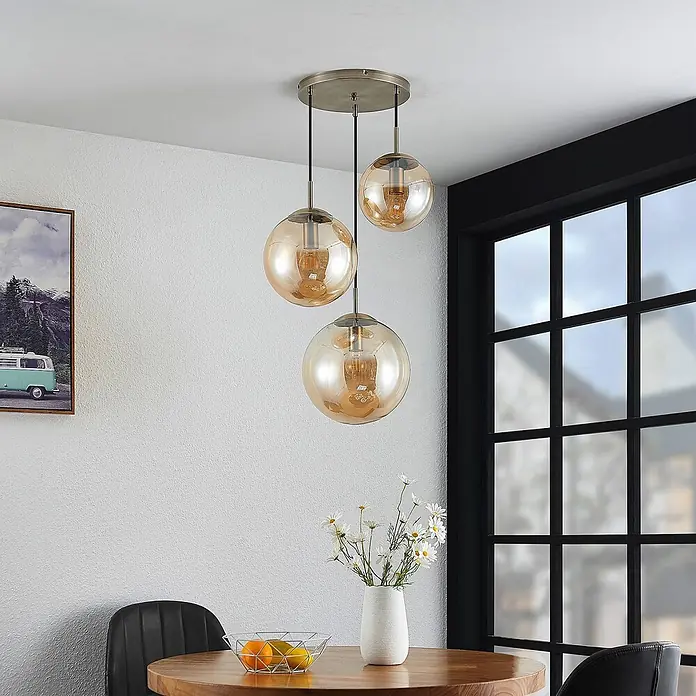 Lindby Pendelleuchte Teeja - creme / amber, amber, altmessing, E27 - Dreiflammige Pendelleuchte mit bernsteinfarbenen Glaskugelschirmen, von einer runden Metallplatte über einem Esstisch hängend.
