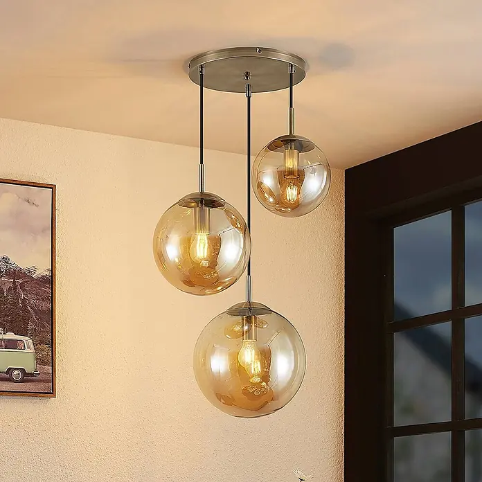 Lindby Pendelleuchte Teeja - creme / amber, amber, altmessing, E27 - Drei bernsteinfarbene Glaskugel-Pendelleuchten mit Messingakzenten, von einer Deckenplatte hängend, beleuchten einen Raum.