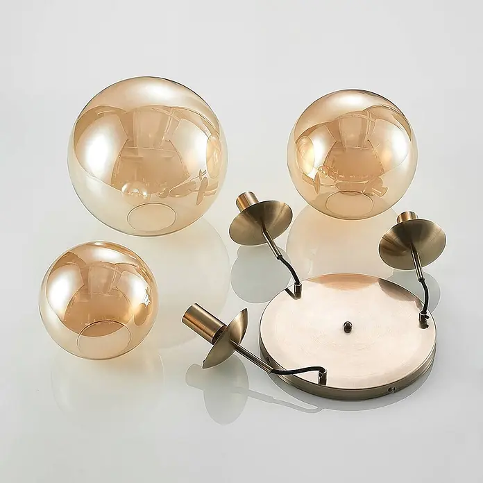 Lindby Pendelleuchte Teeja - creme / amber, amber, altmessing, E27 - Drei bernsteinfarbene Glaskugeln und eine messingfarbene Metallbasis mit Lampenfassungen für eine Deckenleuchte.