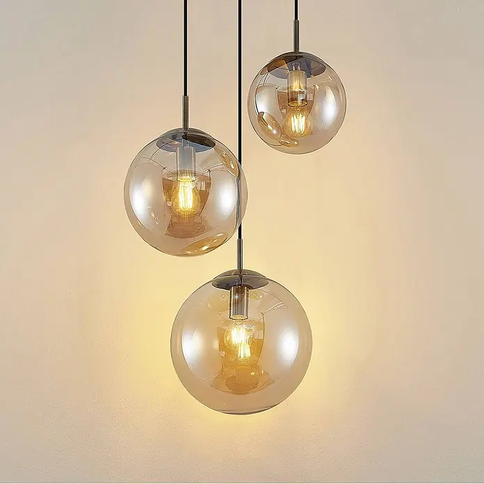 Lindby Pendelleuchte Teeja - creme / amber, amber, altmessing, E27 - Drei Pendelleuchten mit kugelförmigen, bernsteinfarbenen Glasschirmen, schwarzen Kabeln und Metallsockeln, warm beleuchtet.