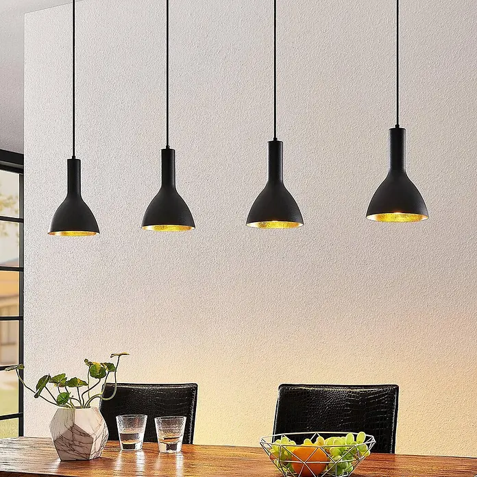 Arcchio Pendelleuchte Cosmina - schwarz, Schwarz, gold, E27 - Vier schwarze Pendelleuchten mit goldener Innenseite hängen in einer Reihe über einem Holztisch und beleuchten ihn warm.