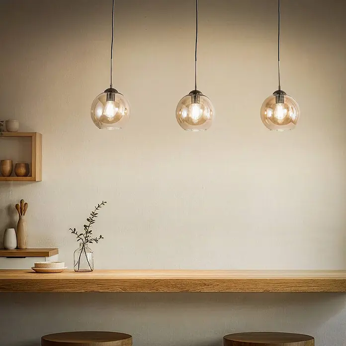 Lindby Pendelleuchte Sofian - L x B x H: 100 x 20 x 20 cm, creme / amber, amber, altmessing, E27 - Drei Pendelleuchten aus Bernsteinglas hängen über einer Holztheke und werfen ein warmes Licht.