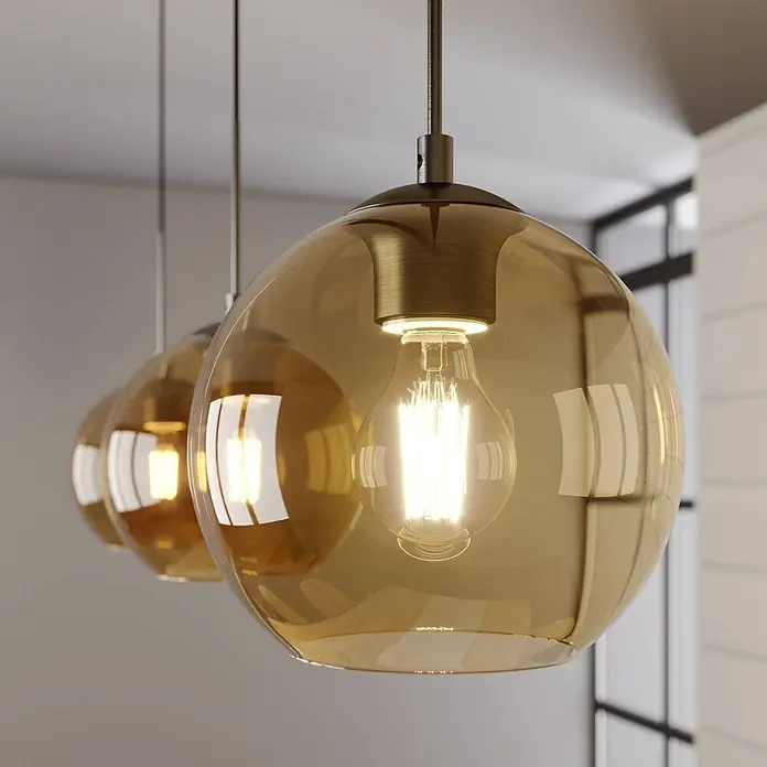 Lindby Pendelleuchte Sofian - L x B x H: 100 x 20 x 20 cm, creme / amber, amber, altmessing, E27 - Drei Pendelleuchten mit bernsteinfarbenen Glaskugeln, Messingbeschlägen und Filamentlampen, von der Decke hängend.