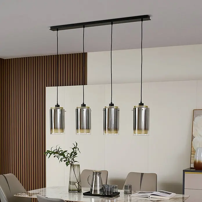 Lucande Pendelleuchte Sterzy - L x B x H: 105 x 15 x 28,5 cm, alu / grau / zink, anthrazit, gold, E27 - Vier moderne Pendelleuchten mit klaren und metallischen zylindrischen Schirmen und Goldakzenten, von einer schwarzen Stange über einem Esstisch hängend.